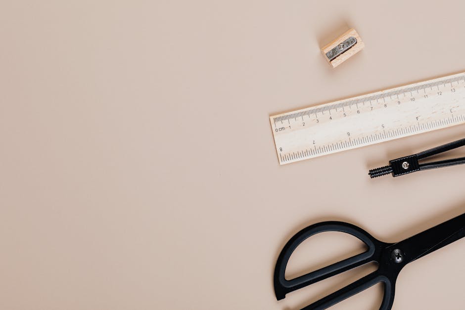 A minimalistic flat lay image featuring a ruler, black scissors, and sharpener on a beige background.