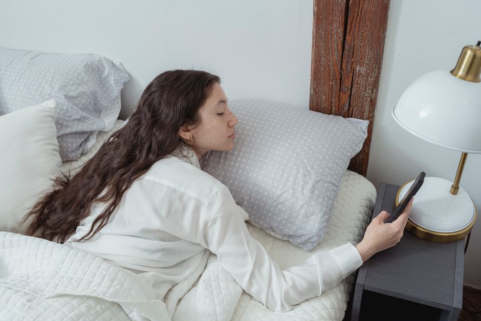 Side view of young brunette with long hair in white nightwear lying on belly in comfortable bed under blanket and using mobile phone in morning at home