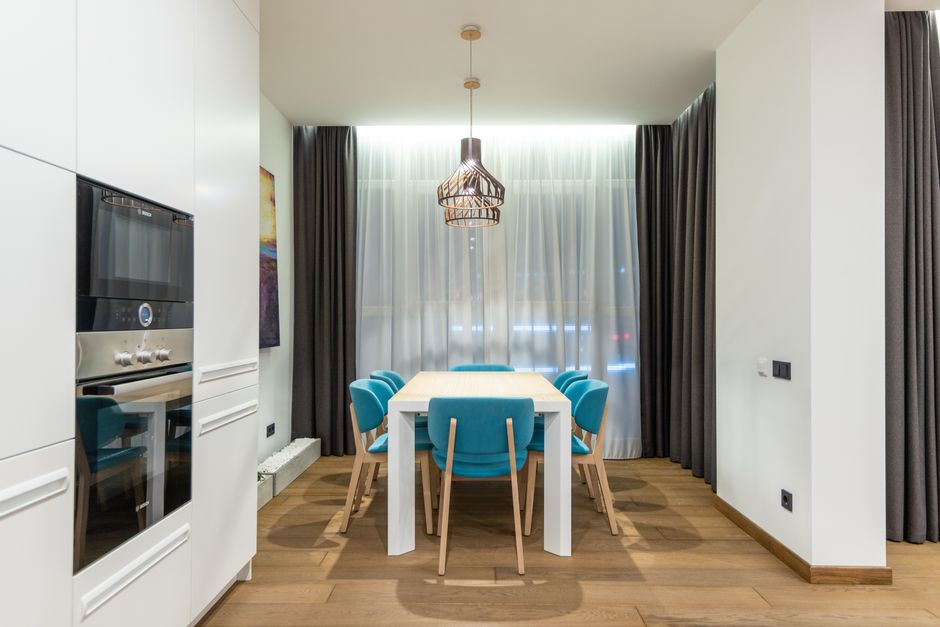 Table with set of turquoise chairs placed at window with curtains in stylish kitchen with white cupboards and modern appliances