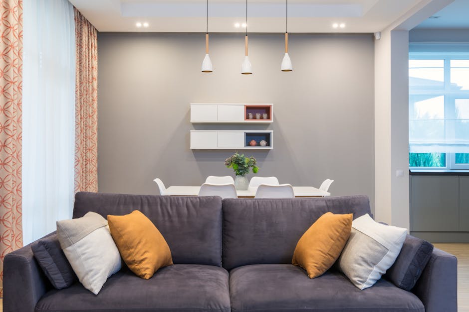Soft sofa with cushions placed near table with chairs at wall with decorations on shelves in spacious flat with lamps