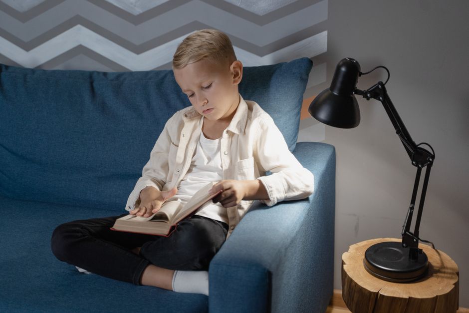 A young boy attentively reading a book while sitting on a cozy blue couch indoors next to a lamp.
