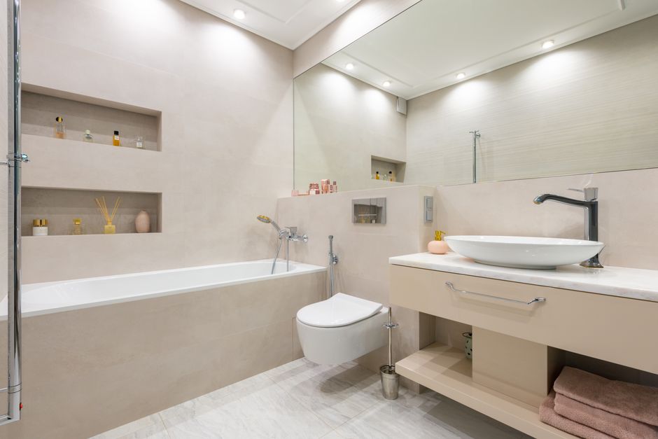 Interior of light modern bathroom with sink and faucet on cabinet with decorative objects next to bathtub and toilet under mirror and shelves