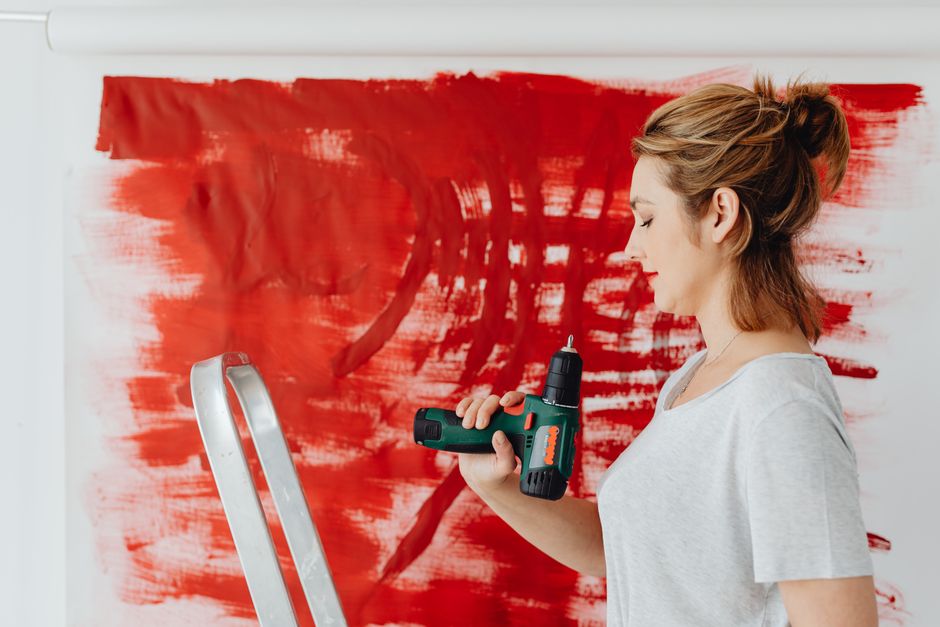 A focused woman using a hand drill near a bright red abstract painting in a studio.
